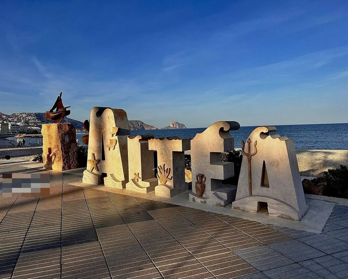 Grandes letras de piedra clara al borde del paseo que forman la palabra ALTEA, cada una con relieves; a la izquierda, sobre un bloque de piedra, una hélice oscura, y detrás el mar al atardecer