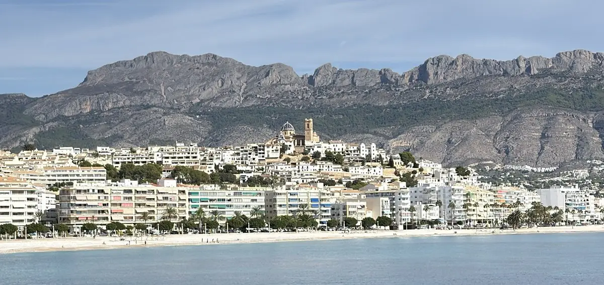 Vista desde el mar hacia Altea: junto a la playa, bloques claros de viviendas y palmeras; detrás, el casco antiguo blanco sube por una colina coronada por la iglesia con cúpula y campanario. Al fondo, una sierra rocosa; cielo algo nublado.
