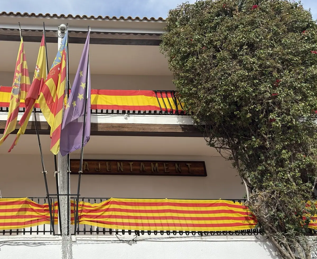 Plantas superiores del ayuntamiento: galerías con vigas de madera, telas a rayas rojas y amarillas en las barandillas y cuatro banderas en mástiles. En la pared, el rótulo de madera «AJUNTAMENT»; a la derecha, una frondosa buganvilla; cielo azul.