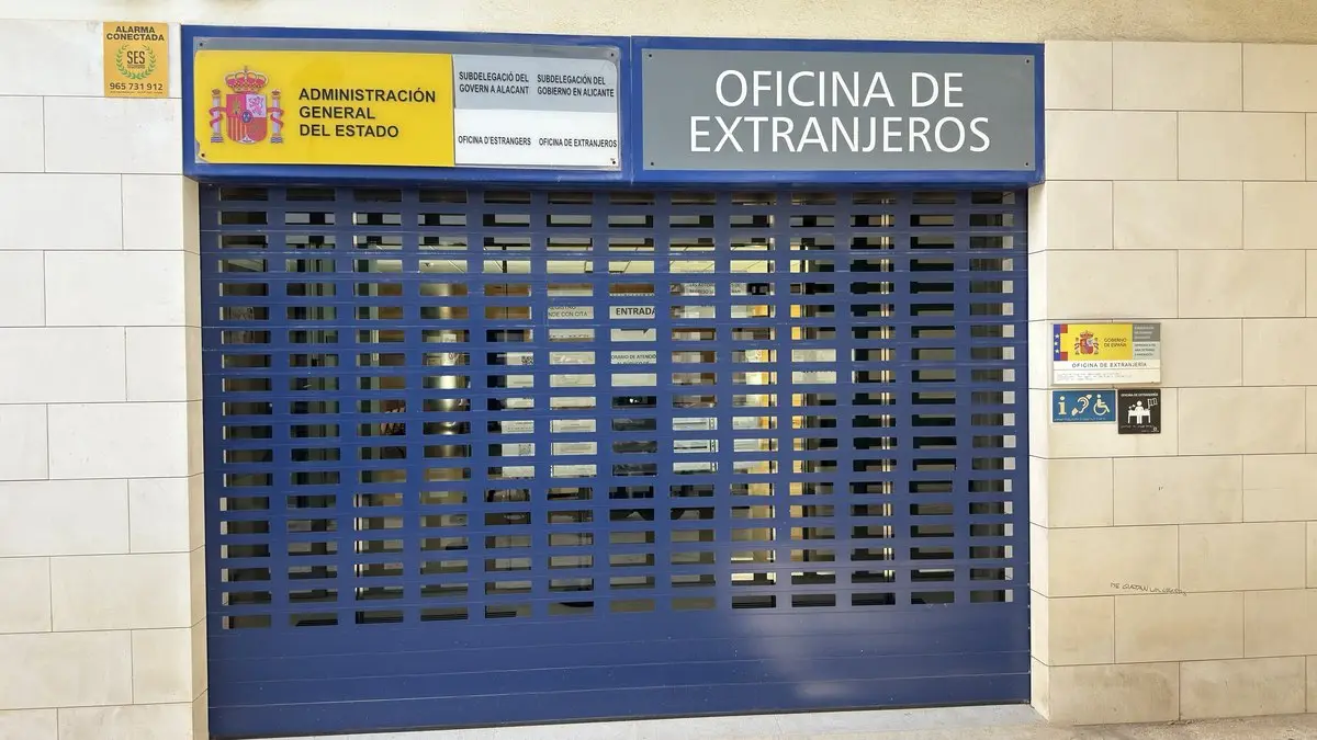 Persiana metálica azul cerrada de una oficina en una fachada de piedra clara. Encima, los rótulos «Administración General del Estado – Subdelegación del Gobierno en Alicante», con el escudo de España, y «Oficina de Extranjeros»; a la derecha, placas menores.