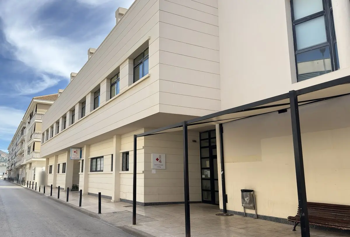 Edificio moderno de color crema, con una hilera de ventanas y entrada cubierta sobre pilares de acero negro; en la pared, los rótulos «Centre de Salut d’Altea» y «Urgències». Delante, una calle con bolardos; cielo con nubes.