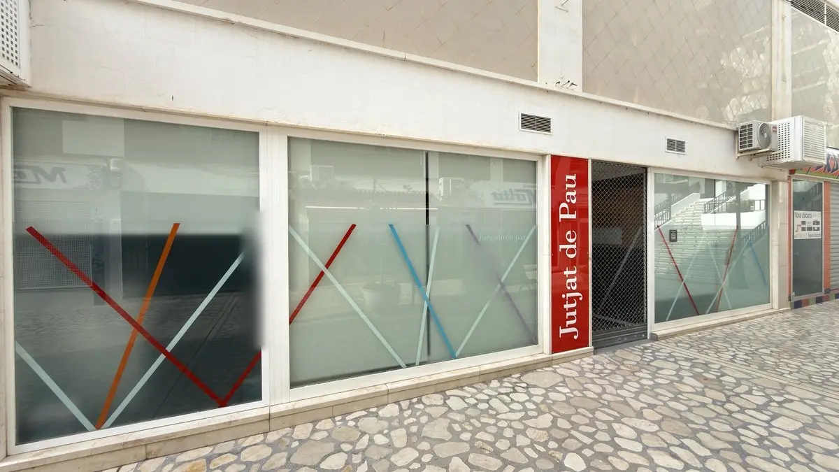 Oficina en planta baja con grandes cristales esmerilados con franjas diagonales de colores y el texto «Juzgado de paz»; al lado, un rótulo rojo vertical «Jutjat de Pau» y una entrada con reja. Delante, una acera de losas irregulares claras.