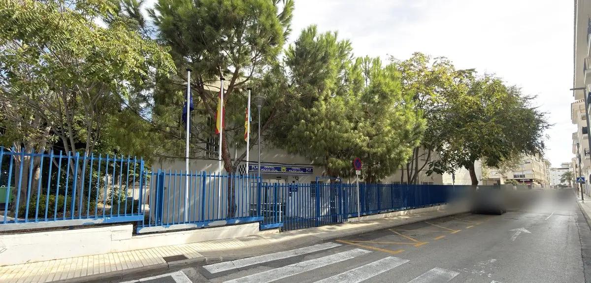 Edificio en esquina tras una valla metálica azul, sombreado por grandes pinos y otros árboles; sobre la puerta, el rótulo «Central Policia Local» y tres mástiles con banderas. Delante, una calle con marcas amarillas.