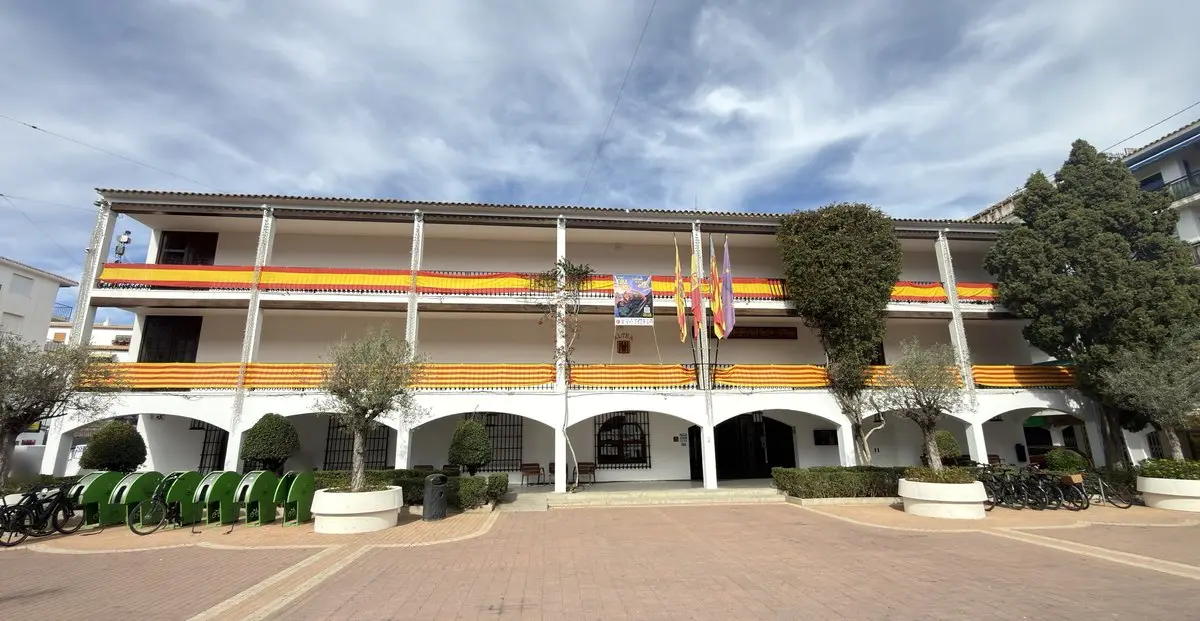 Amplia fachada blanca del ayuntamiento, con dos galerías y soportales; de las barandillas cuelgan telas a rayas rojas y amarillas, y en el centro hay banderas y un cartel. Delante, una plaza con aparcabicis, árboles y maceteros.