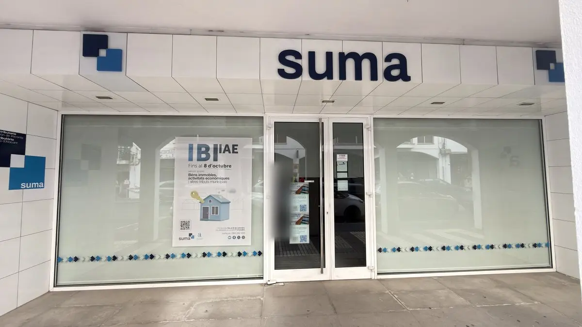 Entrada de oficina con revestimiento de azulejo blanco y el gran rótulo azul oscuro «suma» con su logotipo de cubos. Debajo, una puerta de cristal con avisos y ventanales esmerilados con una franja de rombos azules; en el vidrio se reflejan soportales.