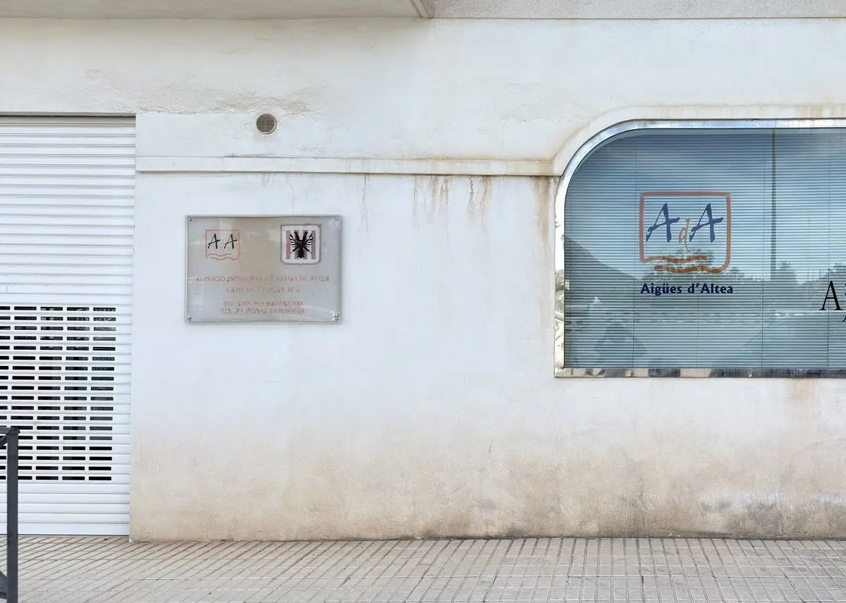 Pared clara y desgastada con una persiana metálica a la izquierda y una placa «Servicio Municipal de Aguas de Altea – Camí de l’Algar nº 6» con teléfonos. A la derecha, una ventana redondeada con estor y el logotipo «Aigües d’Altea».