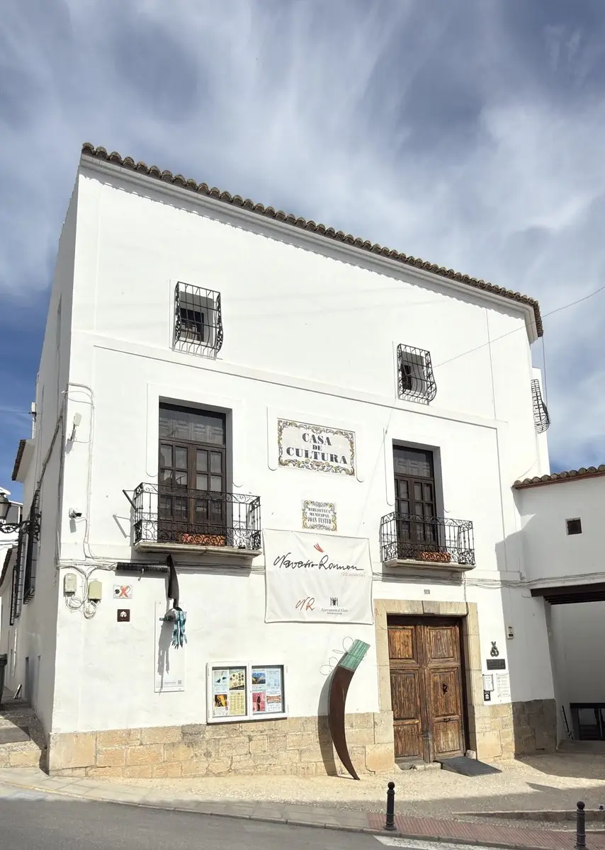 Casa blanca de tres plantas con alero de teja, balcones de forja y antigua puerta de madera enmarcada en piedra. En la fachada, rótulos de azulejo «Casa de Cultura» y «Biblioteca Municipal Joan Fuster», una pancarta «Navarro Ramon» y una escultura metálica curva.