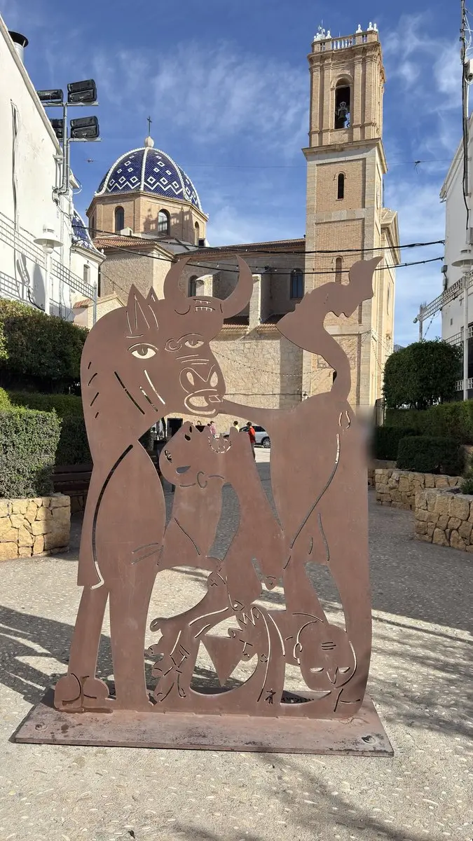 Escultura plana de acero oxidado, recortada como una silueta, en una plaza: un toro con grandes ojos y otras figuras estilizadas. Detrás, la iglesia con cúpula de azulejos azules y blancos y alto campanario; cielo azul, sol.