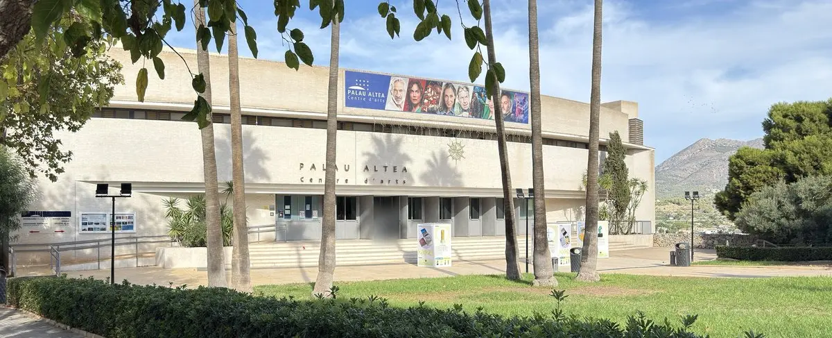 Edificio cultural claro y de líneas rectas con la inscripción «PALAU ALTEA Centre d’arts» sobre la entrada acristalada y una larga pancarta bajo la cornisa. Delante, troncos de palmera, césped y un seto; a la derecha, una montaña; luz solar.