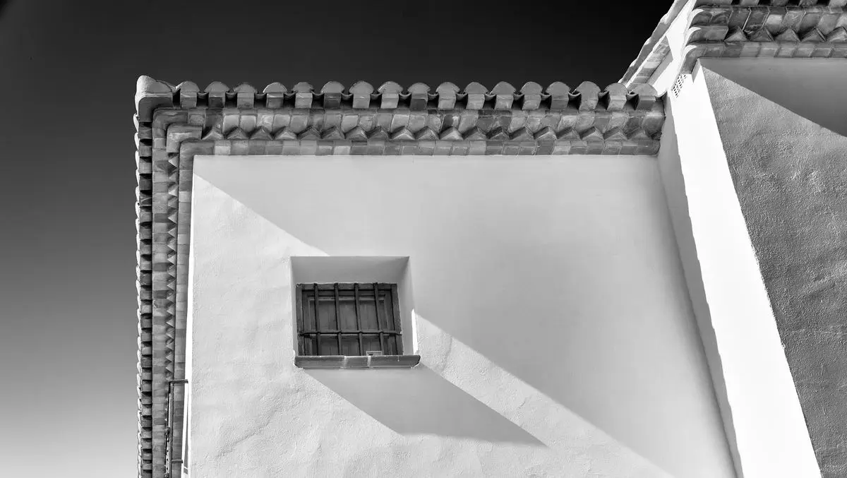 Fotografía en blanco y negro tomada desde abajo contra la esquina de una casa: varias hiladas de teja forman una cornisa dentada bajo el alero; debajo, una ventana pequeña enrejada con postigo de madera y una sombra dura cruzando la pared encalada