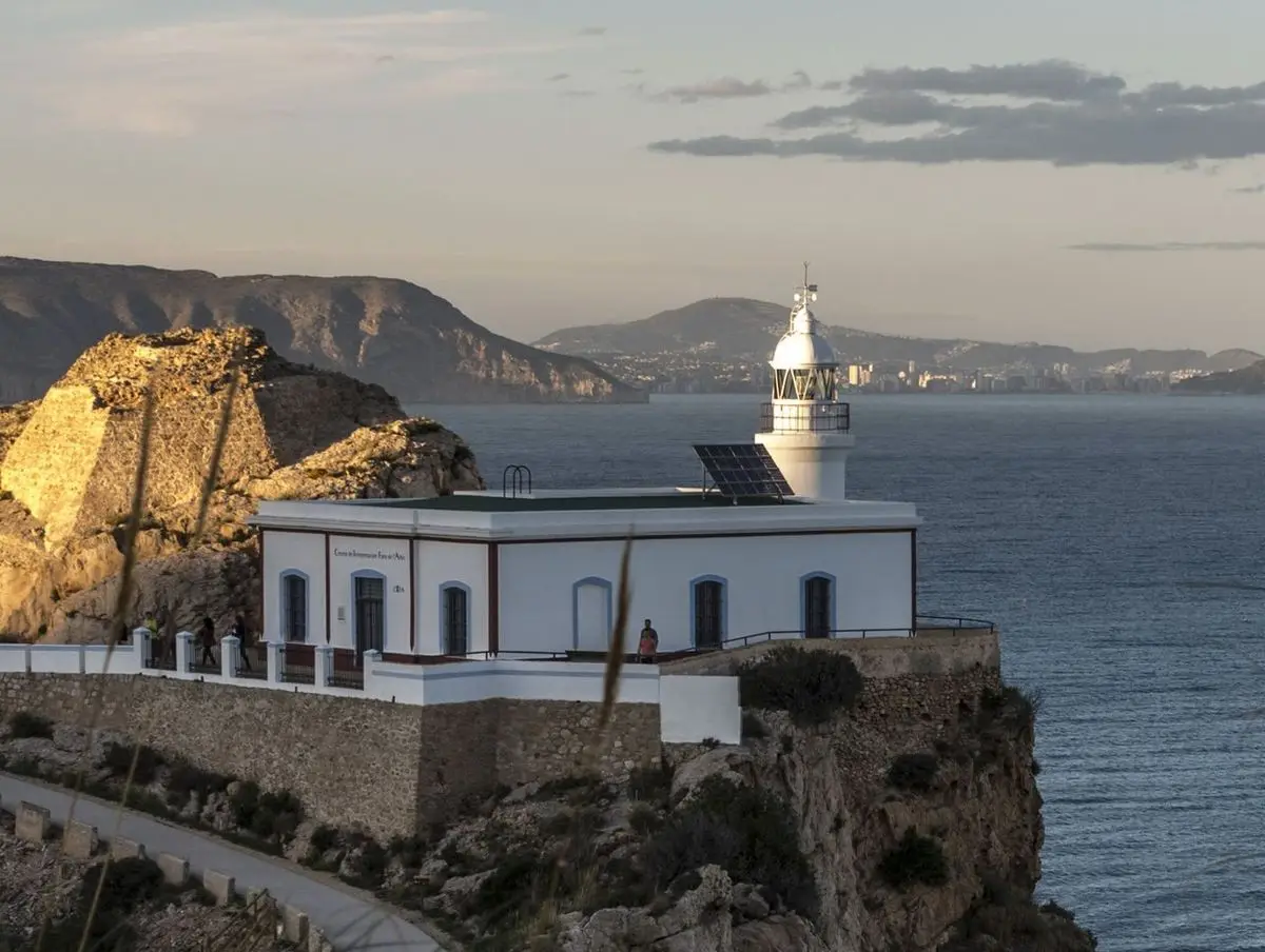 El faro blanco sobre el acantilado a la luz de la tarde, delante la casa baja del farero y, al fondo, la bahía y las montañas
