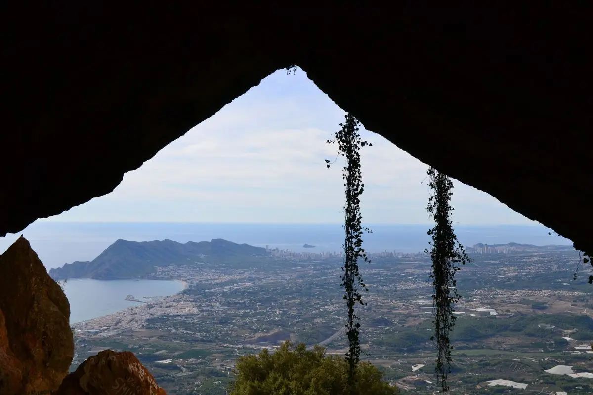 Vista desde un oscuro paso en la roca hacia la costa, muy abajo: la bahía, el pueblo y los rascacielos al fondo; en el marco, hiedra colgante