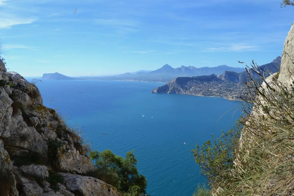 Vista desde lo alto de una roca a lo largo de la costa: abajo el mar azul intenso, a la derecha un cabo densamente edificado y, detrás, una amplia bahía y montañas entre la calima