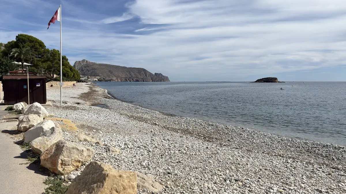 Amplia playa de cantos claros con grandes rocas junto a un paseo; a la derecha, mar en calma con un pequeño islote. A la izquierda, una caseta de madera, un mástil con bandera granate y árboles; al fondo, una montaña junto al mar.