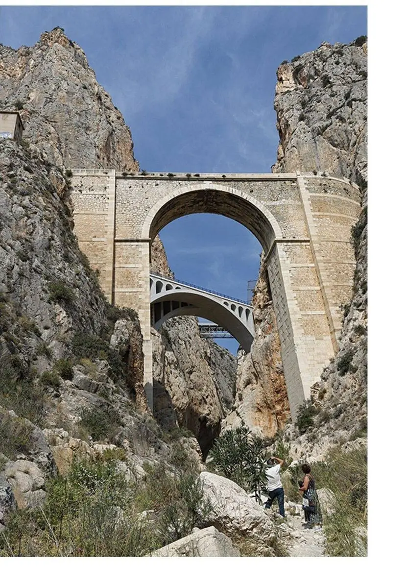 Vista hacia arriba desde el lecho de un barranco: un arco de piedra entre altas paredes de roca y, a través del arco, un segundo puente en arco y un puente metálico; abajo, dos personas, una de ellas fotografiando