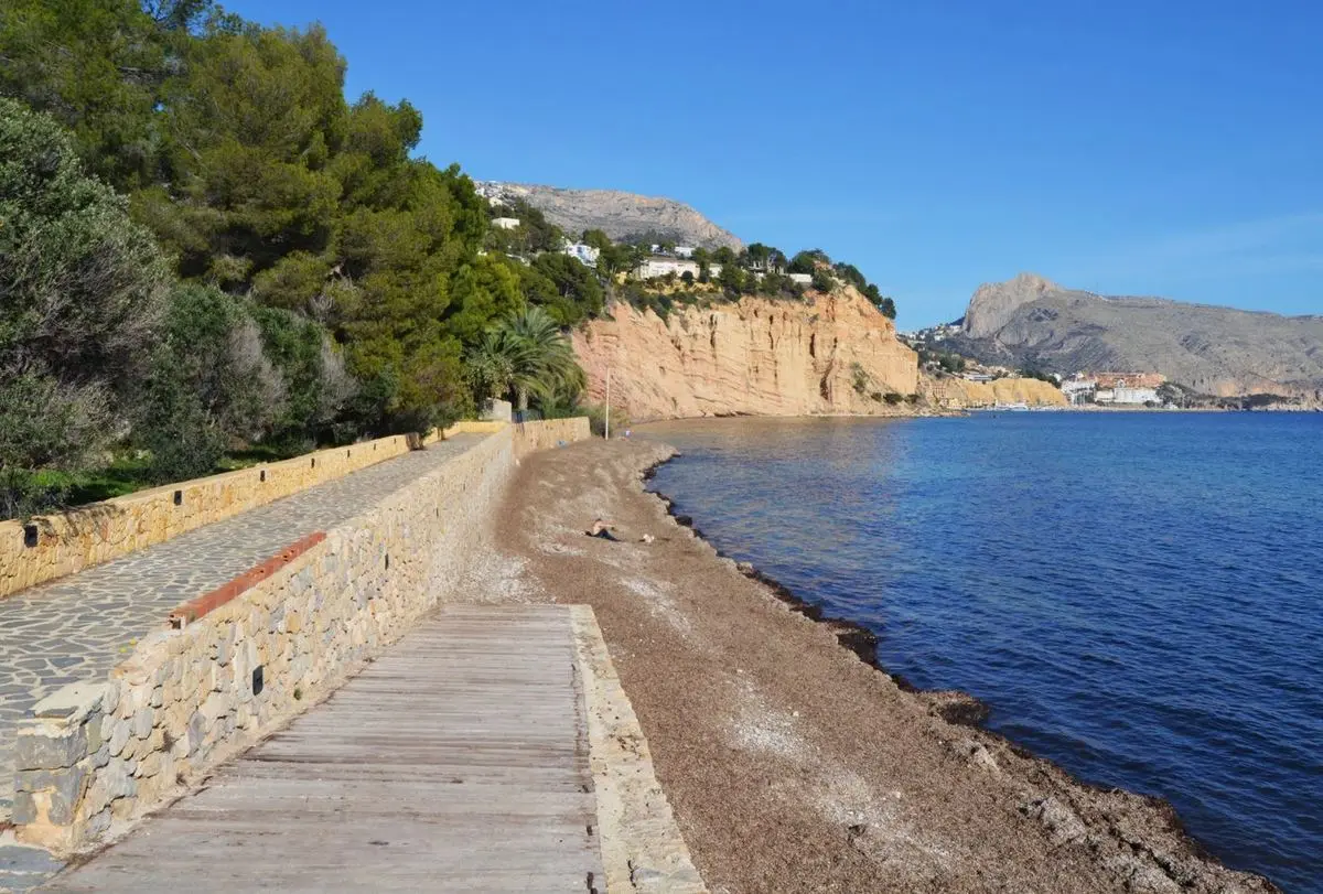 Un camino empedrado tras un muro de piedra junto a la playa de cantos, a la derecha el mar y al fondo un acantilado ocre