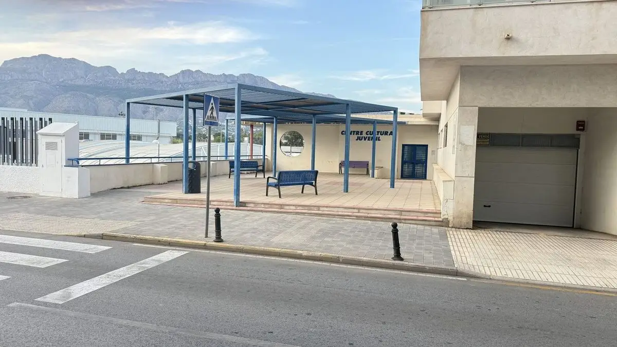 Een laag, licht gebouw met het opschrift Centre Cultural Juvenil, ervoor een blauwe overkapping op pijlers met banken eronder, en op de achtergrond de steile wand van de Serra de Bèrnia