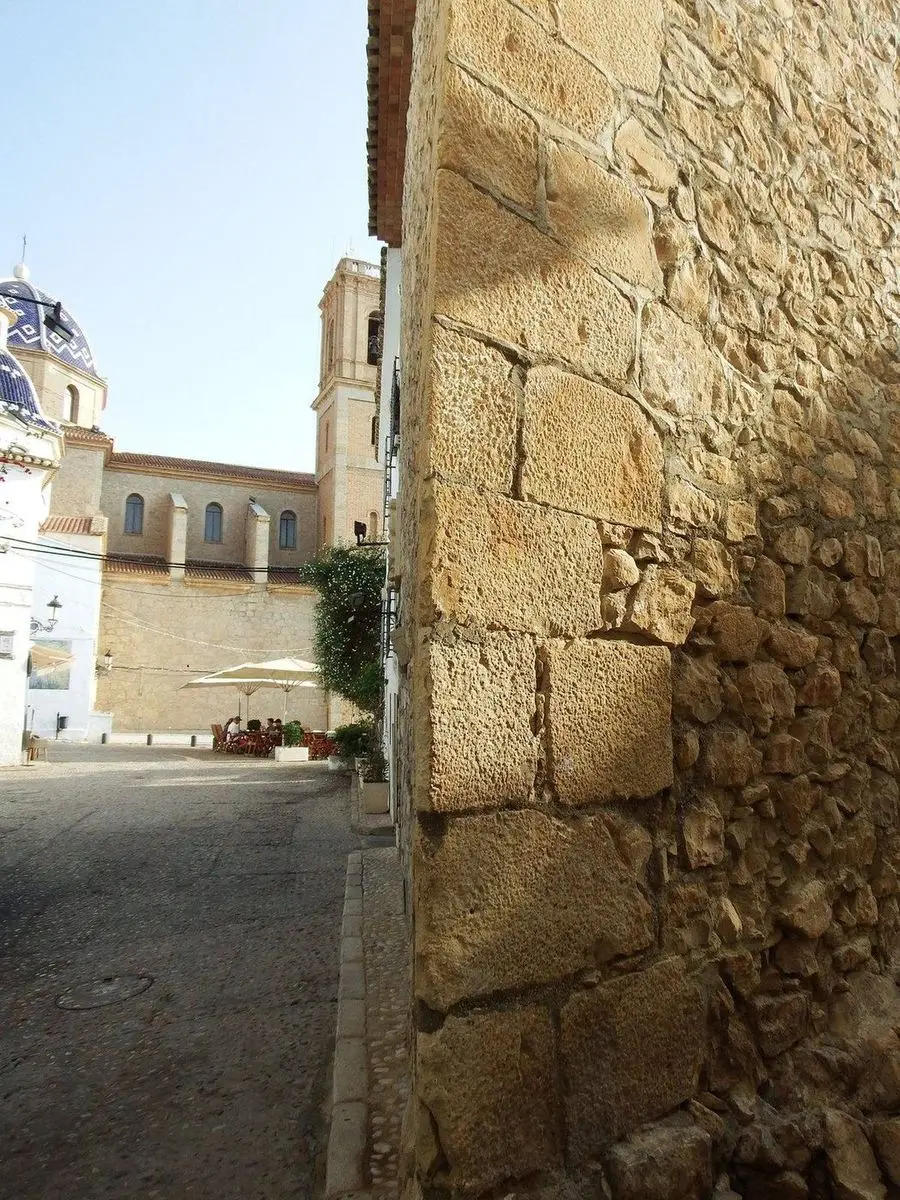 Un muro de grandes sillares y mampostería en primer término y, detrás, la plaza de la iglesia con la cúpula azul y el campanario