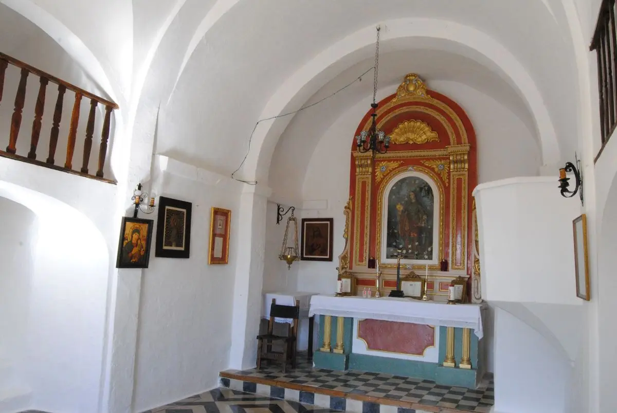 El interior encalado de una pequeña ermita: dos bóvedas de cañón de radios distintos, a la izquierda una tribuna elevada con barandilla de madera torneada, cuadros enmarcados en las paredes, sobre el altar un retablo rojo y dorado con la imagen de la santa, el suelo de baldosa blanca y negra