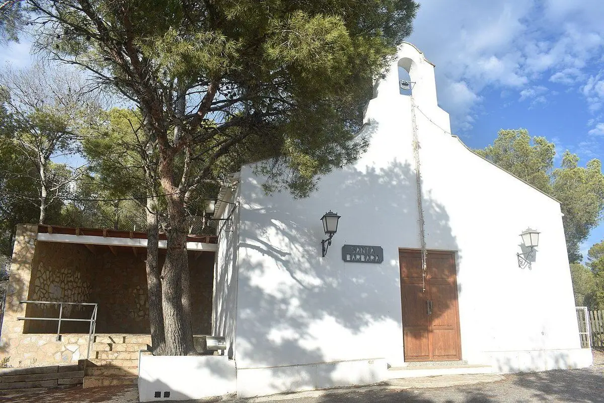 La ermita de Santa Bàrbara, con espadaña, rótulo y el porche cubierto a la izquierda, y un pino delante