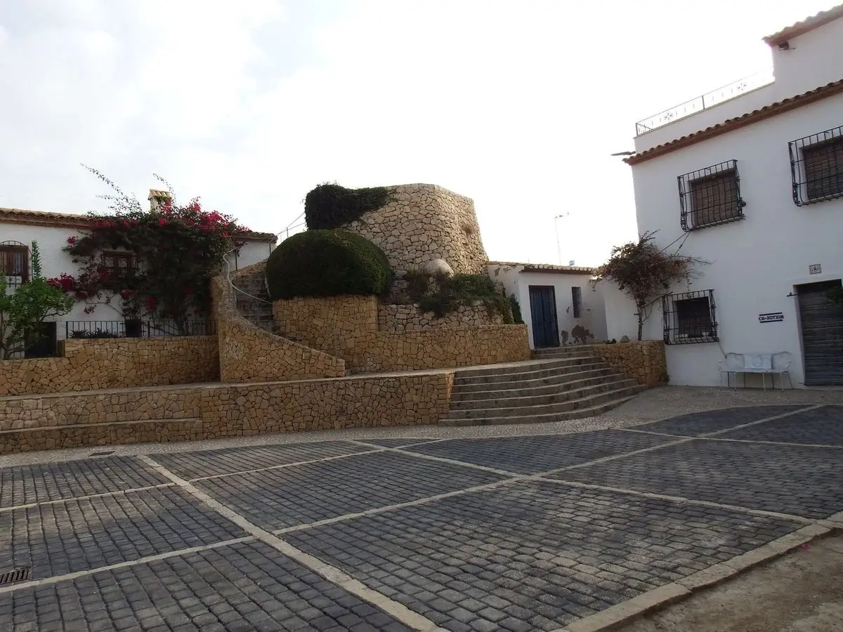 La placeta empedrada ante la torre: el muñón de mampostería sobre bancales de piedra seca, a la izquierda una casa con buganvilla y a la derecha una fachada blanca