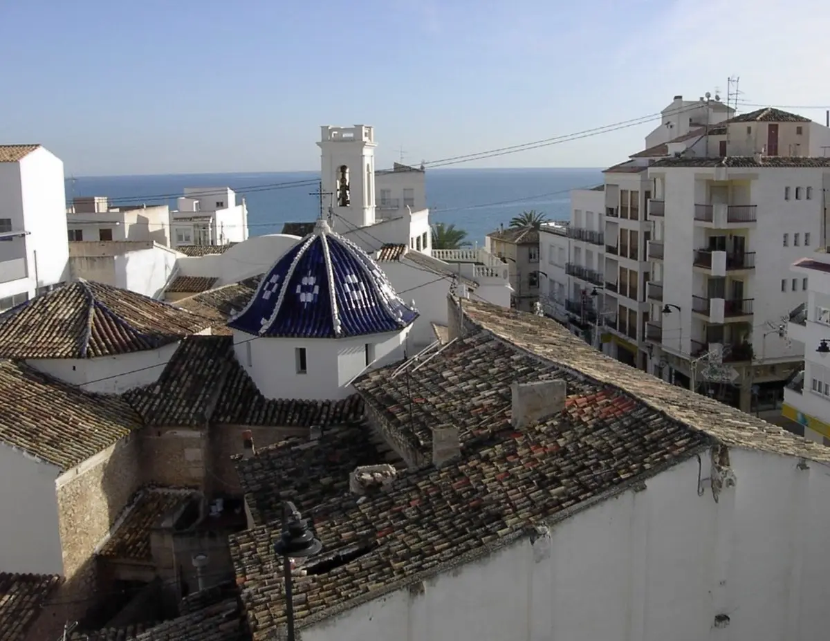 Vista sobre viejos tejados hacia una pequeña cúpula octogonal de tejas azul oscuro con rombos blancos y, detrás, un campanario encalado con una campana; a la derecha, bloques de viviendas con balcones y, en el horizonte, el mar.