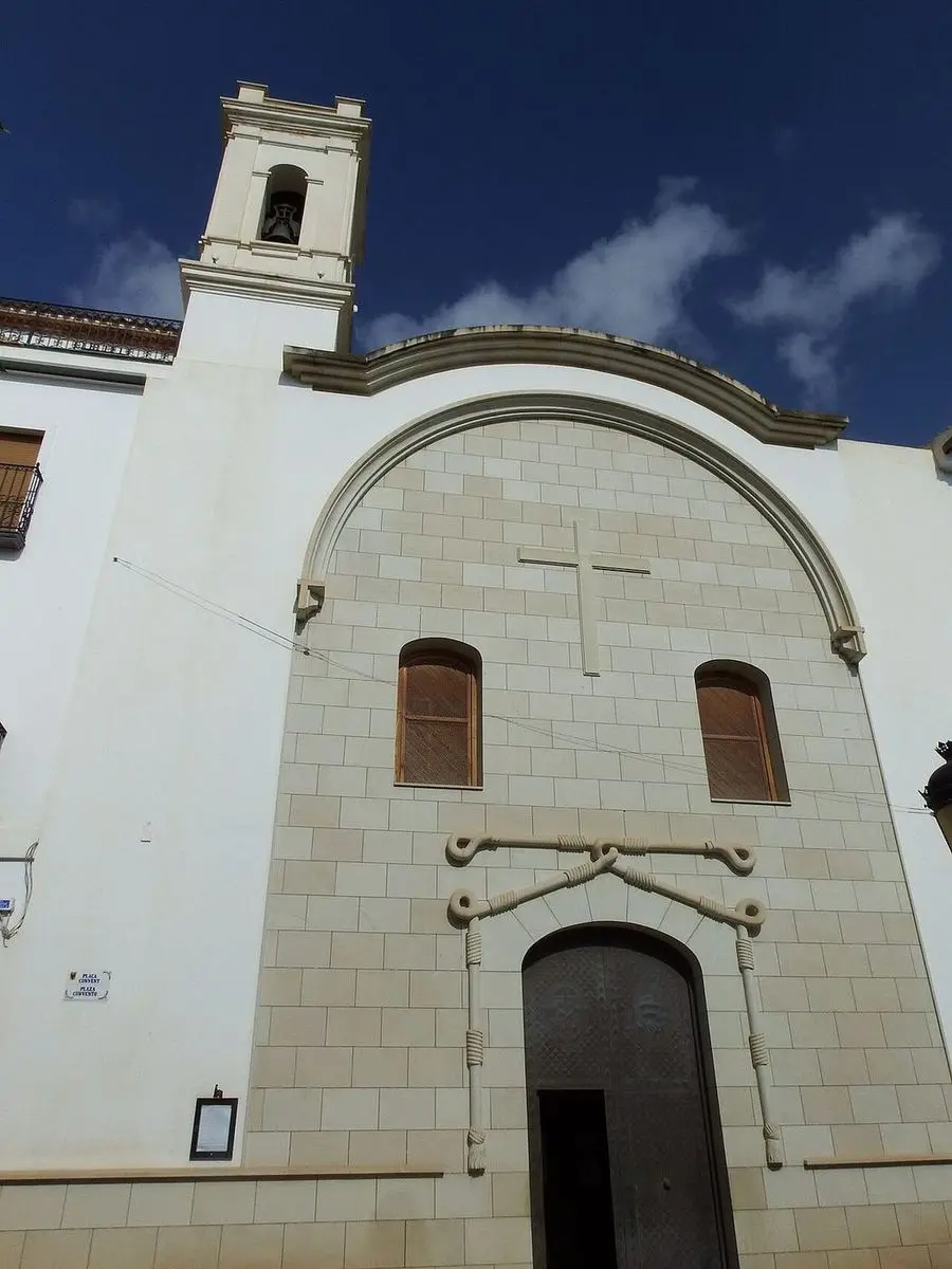 La fachada de la iglesia franciscana de cerca: cruz en el tímpano, dos ventanas de medio punto y, sobre la portada, un cordón de piedra con nudos