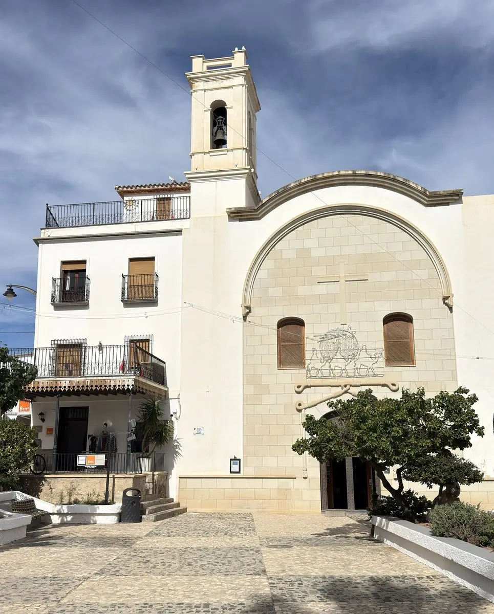 Fachada blanca de iglesia con pequeño campanario de una sola campana; en el gran arco de sillares claros, una cruz y un relieve, y debajo la entrada tras un árbol. A la izquierda, una casa con balcones; delante, una plaza empedrada.