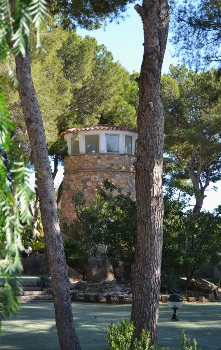 Una torre redonda de mampostería y canto rodado entre pinos, con el cuerpo superior acristalado y cubierta cónica de teja; delante, un jardín con escalones, pitas y un farol esférico