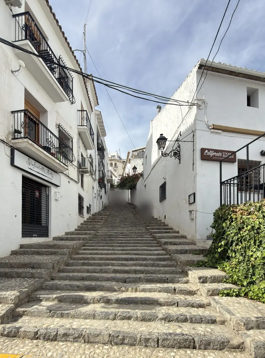 Una amplia escalinata de peldaños bajos de piedra y canto rodado sube entre casas blancas con balcones de hierro; a la derecha, hiedra y un farol de forja; arriba, buganvilla en flor y algunos peatones.