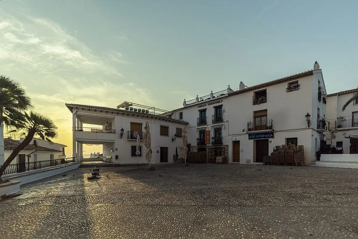 La plaza de la iglesia vacía con la primera luz: empedrado de cantos, casas blancas con balcones y, por un arco a la izquierda, el cielo claro sobre el mar