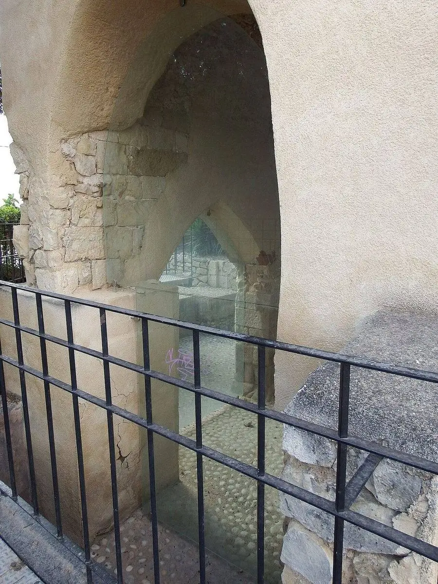 Un arco apuntado de mampostería tras un vidrio y una reja de hierro, y detrás más obra antigua