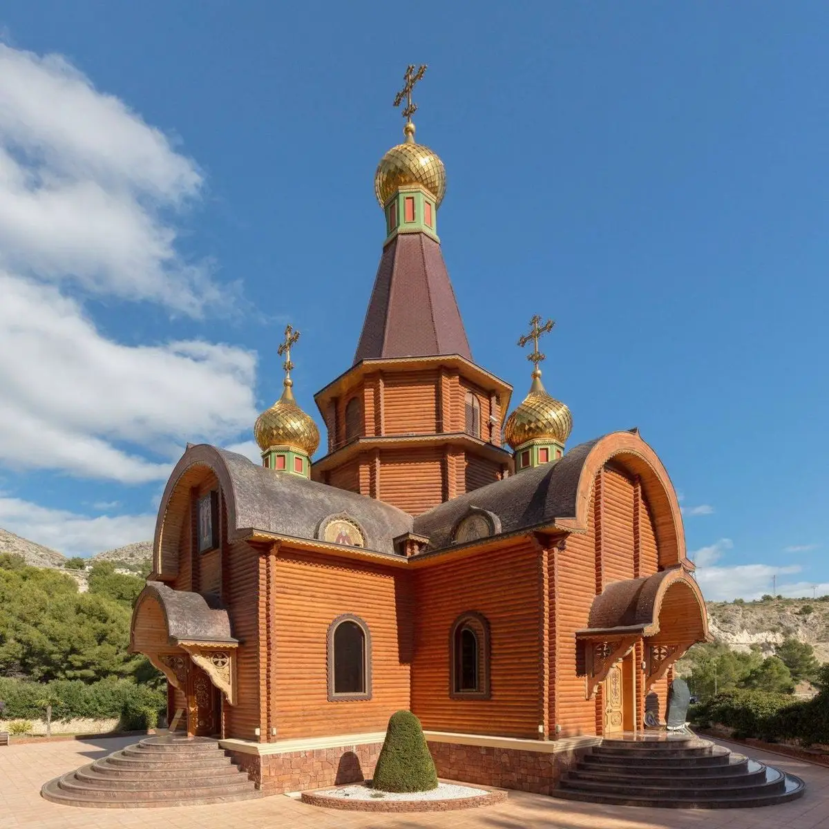 La iglesia de madera de frente: muros de troncos encajados, tres cúpulas de bulbo doradas y una torre octogonal, con escalinatas de piedra alrededor
