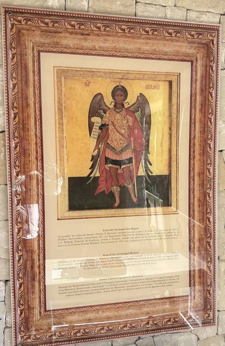Icono enmarcado tras un cristal en una pared de piedra: el arcángel Miguel con alas, armadura, manto rojo y una filacteria sobre fondo dorado; debajo, una cartela en español, inglés y ruso.