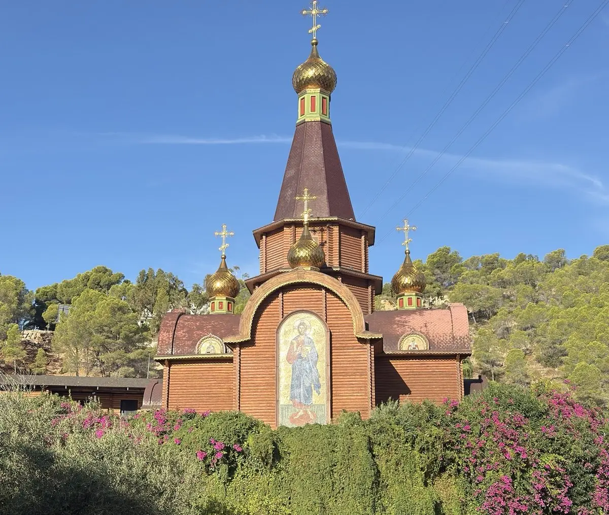 Iglesia ortodoxa de madera de color pardo rojizo, con torre puntiaguda, cúpulas doradas en forma de bulbo y cruces; en la fachada, un gran mosaico de Cristo de pie. Delante, setos y buganvillas fucsia; detrás, una ladera de pinos bajo cielo azul.