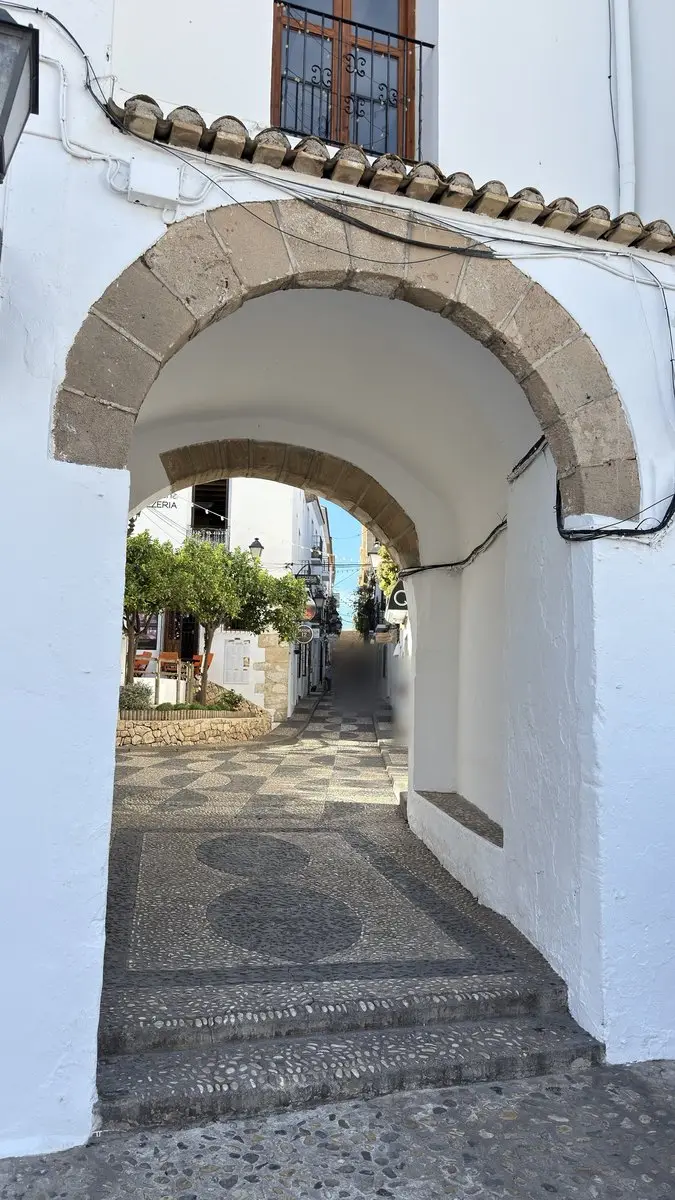 Vista a través de un pasaje encalado con dos arcos de piedra hacia una calle soleada del casco antiguo. Dos escalones de canto rodado suben al pasaje, con suelo de guijarros claros y oscuros; al fondo, árboles, farolas y casas blancas.