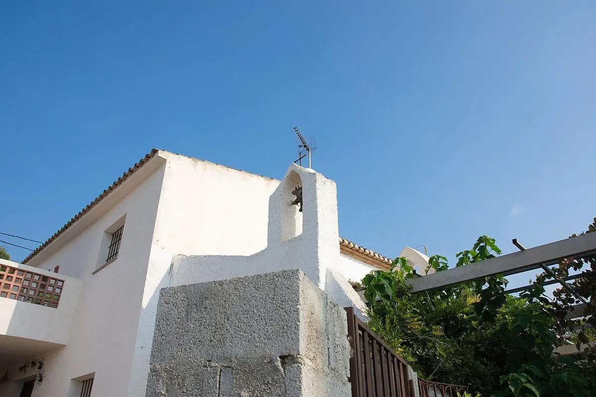 La espadaña de Sant Tomàs sobre el muro blanco, entre viviendas y una parra