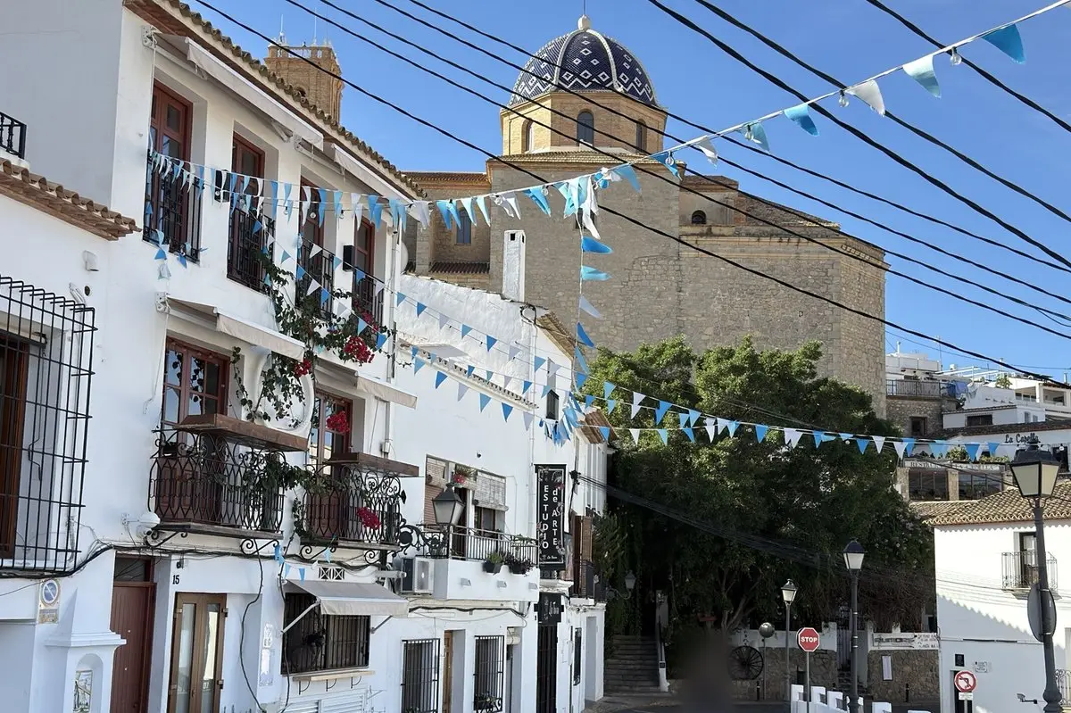 Casas encaladas con balcones de forja y flores junto a una plaza empedrada; guirnaldas de banderines azules y blancos y cables cruzan por encima, detrás árboles y la cúpula de tejas vidriadas de la iglesia.