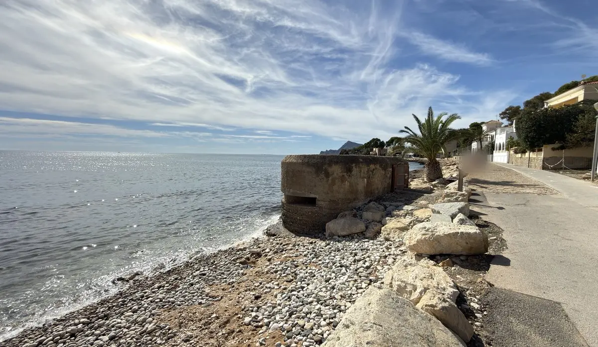 Búnker de hormigón redondeado con aspillera sobre una playa de cantos, rodeado de rocas; a la izquierda, el mar brillando a contraluz, y a la derecha, un paseo junto a villas y una palmera. Al fondo, una montaña costera; cielo con cirros.