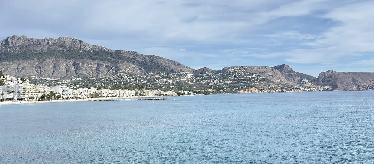 Un mar tranquilo de color turquesa ocupa el primer plano; a la izquierda, una hilera de casas blancas con palmeras en la orilla, detrás montañas peladas y laderas salpicadas de chalés, y a la derecha acantilados.