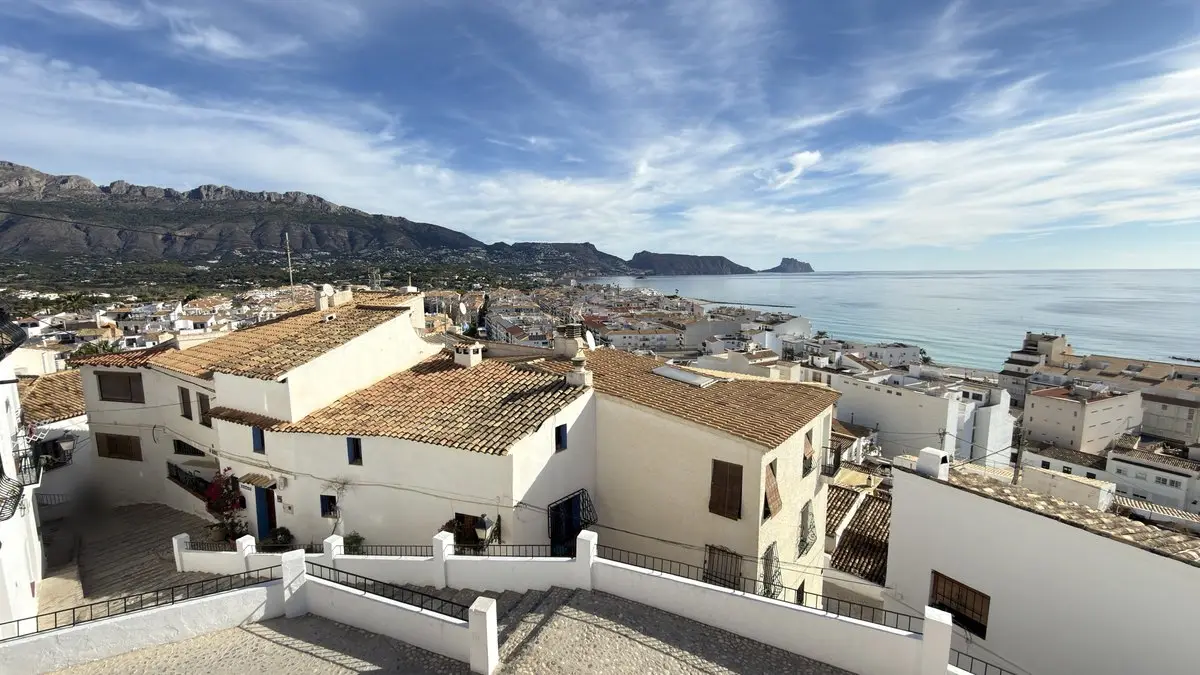 Vista elevada de casas blancas con tejados de teja y una calle escalonada del casco antiguo; detrás, la ciudad hasta la costa, el mar turquesa a la derecha y una sierra que termina en una roca marina. Cielo soleado con nubes tenues.