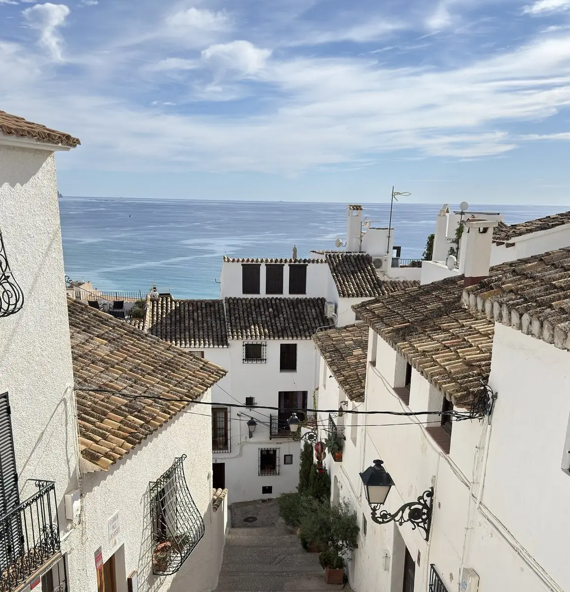 Desde un balcón con barandilla de hierro forjado se ve una calle en pendiente entre casas encaladas con tejados de teja, farolas de pared y macetas; al fondo, el mar azul bajo nubes ligeras.