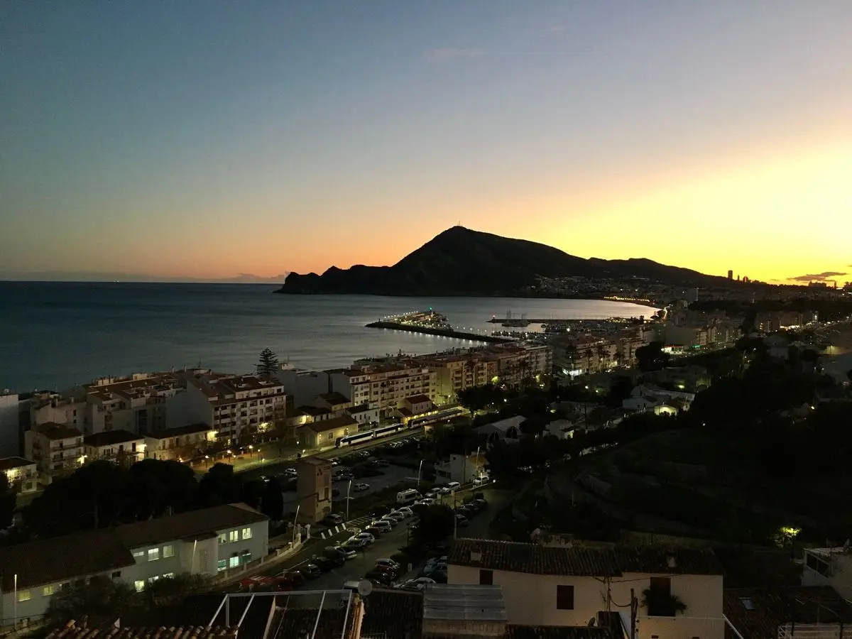 La misma bahía después de la puesta de sol: el cielo todavía naranja sobre la montaña, abajo las calles iluminadas, un tren del TRAM en la parada y las luces del puerto