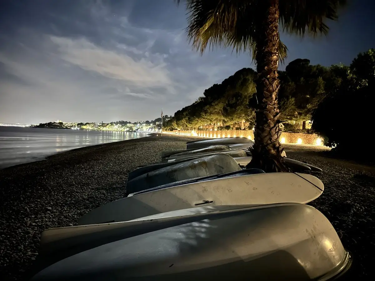 Fotografía nocturna desde la playa de cantos: una hilera de barcas volcadas en primer plano, junto a ellas una palmera iluminada y al fondo la bahía y las luces de Altea bajo un cielo azul oscuro