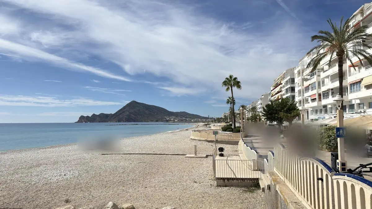 Playa de cantos y paseo marítimo bajo nubes altas; a la derecha, bloques blancos de apartamentos, palmeras y una barandilla curva de pasamanos azul; a la izquierda, el mar en calma y al fondo la Serra Gelada.