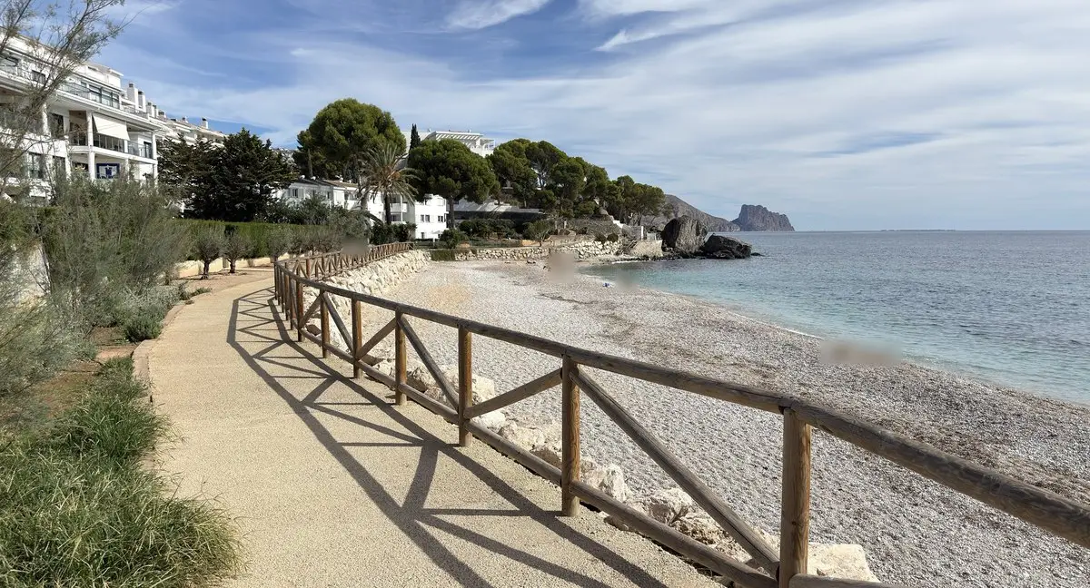 Un sendero de color arena con barandilla de madera bordea una playa de cantos; a la izquierda hierbas, pinos y edificios blancos, a la derecha el mar claro y tranquilo y, a lo lejos, el Penyal d’Ifac.