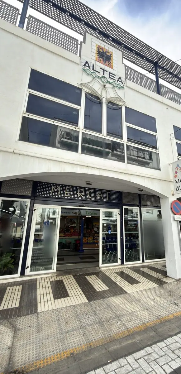 Wit marktgebouw met grote donkere ramen; bovenaan het woord “ALTEA” onder een wapen van tegels met een adelaar en lichtversiering in de vorm van klokken. Op de begane grond glazen deuren onder het opschrift “MERCAT”; ervoor bestrating met strepen.