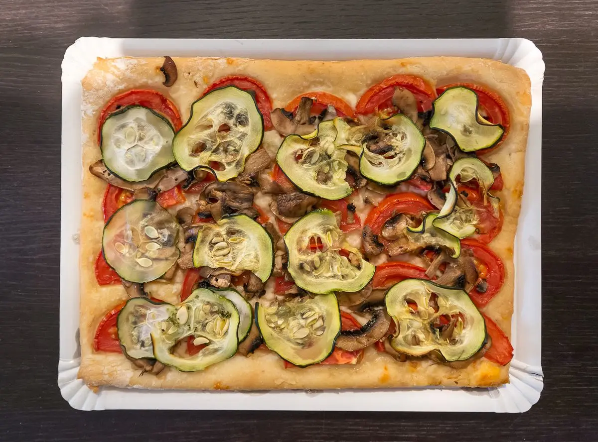 Una coca rectangular de masa de levadura sobre una bandeja de cartón, cubierta de rodajas de tomate, calabacín y champiñones, con los bordes dorados