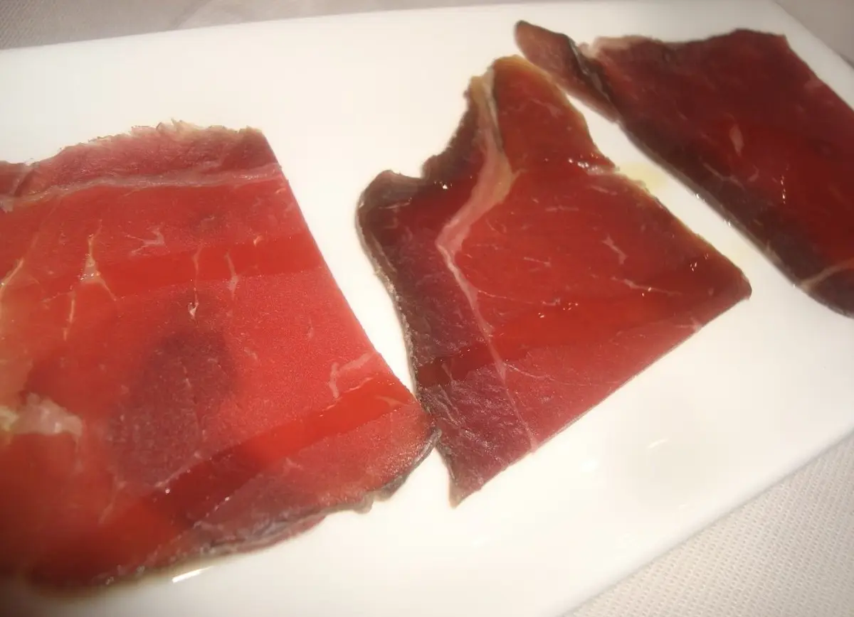 Tres lonchas finas de atún seco, de color rojo oscuro, sobre una bandeja blanca, con brillo de aceite de oliva y vetas claras.