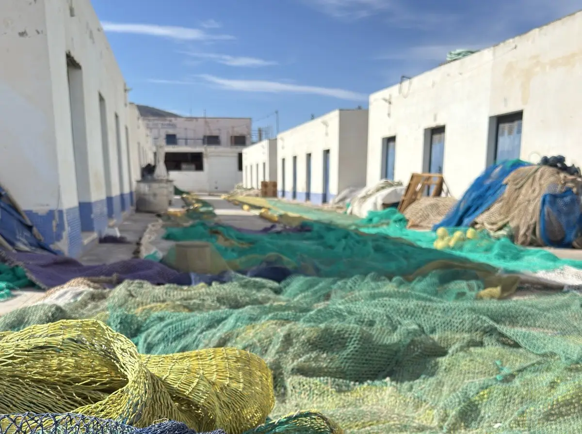 Redes de pesca verdes, amarillas y azules, extendidas y amontonadas en el suelo entre dos hileras de almacenes bajos encalados con puertas y zócalo azules; arriba, cielo azul con nubes finas.